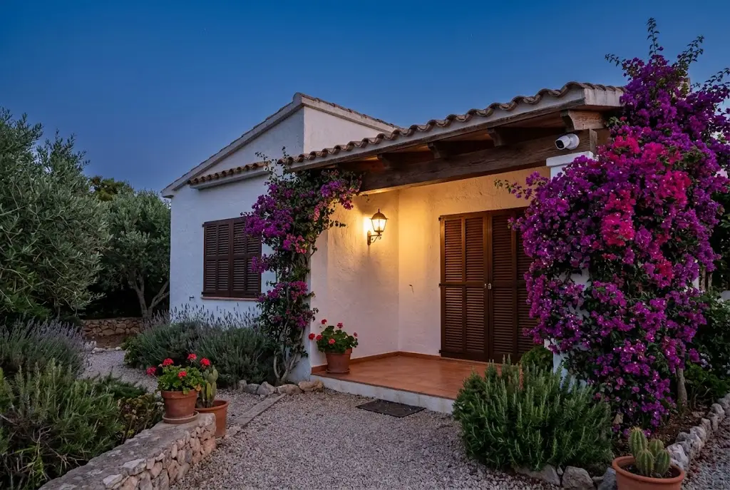 Casa de veraneo mediterránea al anochecer, cerrada y con las persianas bajadas, con la luz del porche encendida y una cámara de vigilancia bajo el alero